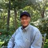 African-American man with glasses wearing a blue cap and button-up shirt sitting outside with trees and plants behind him