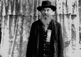 Older white man in hat and suit with long beard standing with curtains behind him