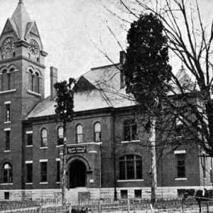 Brick building with clock tower, water tower, iron fence, and horse and carriage, trees