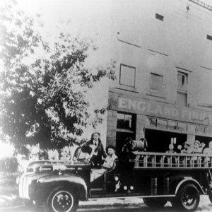 Fire trucks with white firemen parked outside brick building with sign "England Fire Dept"