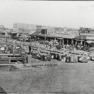 Bales of cotton in town square with row of storefronts on the right with horse drawn wagons in front of them