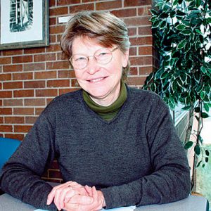 White woman in glasses wearing a sweater sitting at a table with brick wall behind her