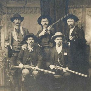 Five white men in western clothing and wide brimmed hats posing with rifles in hand