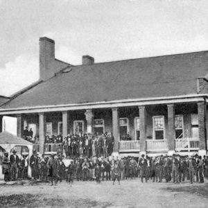 Crowd of white men in suits and hats standing outside of and on steps of two-story brick building with covered porch and gazebo