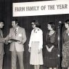 White man in suit handing award to white man in suit standing next to white women in dresses on stage under "Farm Family of the Year" banner