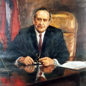 White man in black suit sitting at his desk with Arkansas flag behind him