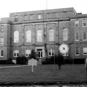 multistory brick building with lawn trees sidewalk and round sign labeled "76' and historical sign