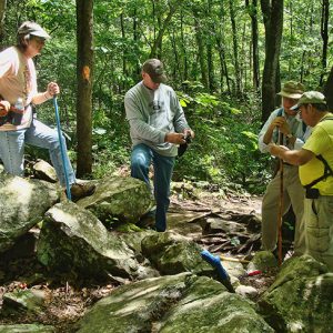 Group of white men and a woman standing on and near rocks in forested area