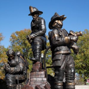 Bronze statue of two firefighters in full gear using hoses while a third firefighter comforts a small child behind them