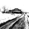 Muddy rutted road with snow alongside leading to brick building with parked cars and bare tree