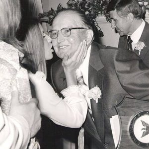 White woman kissing older white man with glasses in suit and tie holding award plaque with white man in suit standing behind him