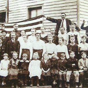 Group of white children and white man holding up an American flag outside school house