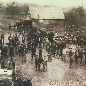 Crowd of people and horse drawn wagons on dirt road with single-story storefronts on both sides