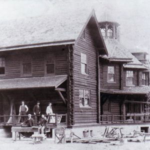 White men standing outside large two-story log structure with tower