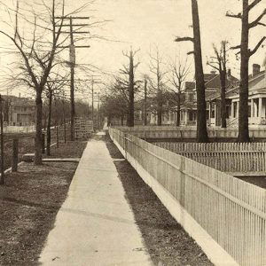 Looking down sidewalk in residential neighborhood with houses inside fences on dirt street