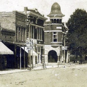 Dirt street with brick storefronts and building with tower and trees