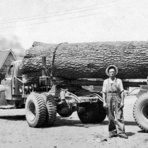 White man in overalls and hat with logging truck carrying a massive log and building with truck in the background