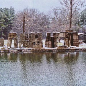 Concrete ruins near edge of pond with pavilion and trees in background during winter