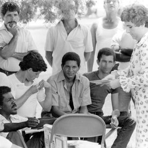 White woman talking to a group of Cuban men standing and sitting under a tree