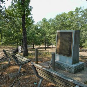 Stone monument with plaque and benches on sloped hill top in forested area