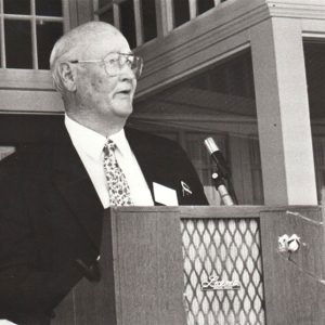 White man in suit and tie speaking into microphone at lectern