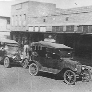 Cars parked outside brick storefronts on dirt road