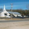 Single-story building with covered porch and steeple with parking lot and sign on street