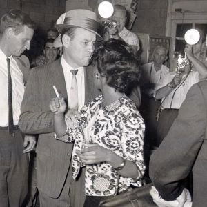 African-American woman being arrested by white men in suits with white photographers taking pictures behind them