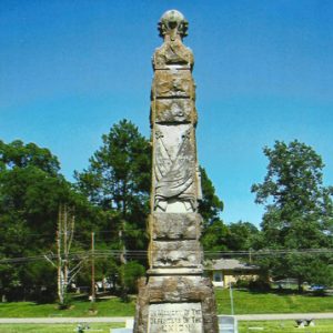 Tall monument with engraving in cemetery with green grass and green trees in background