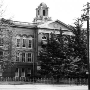 Multistory brick building with classical facade arched windows domed tower and trees behind iron fence