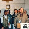 White man in suit speaking at a lectern with mixed group of men and women behind him