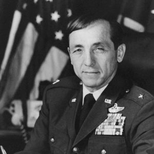 White man in military uniform with flag behind him