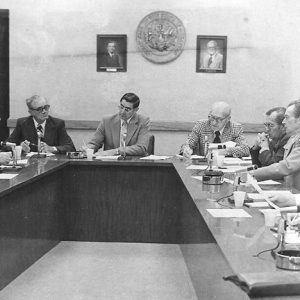 Group of older white men in suits sitting at u-shaped table with microphones in committee room with seal on the wall