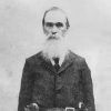 Older white man with long white beard standing in suit with hoster belt over his coat