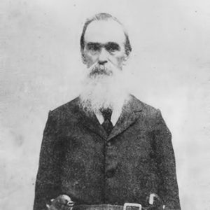 Older white man with long white beard standing in suit with hoster belt over his coat