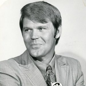 White man with short hair in suit and tie in front of a microphone