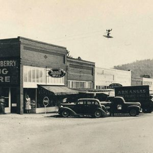 Cars and trucks parked outside row of brick storefronts with tree-covered hill in the background