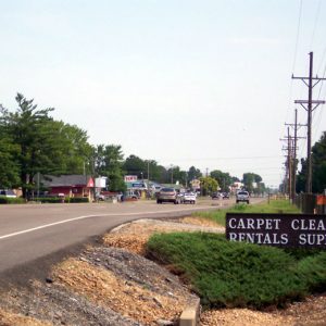 Street with cars and buildings on the left and "Carpet Cleaning Rental Supplies" sign next to a line of telephone polls on the right