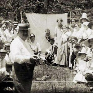 Crowd of white men and women gathered together with man putting his head through a sheet in the background