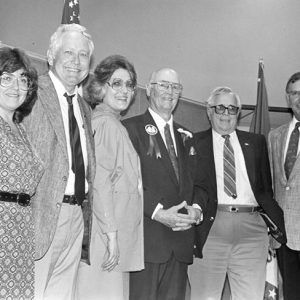 White men and women standing and smiling in line with flags behind them