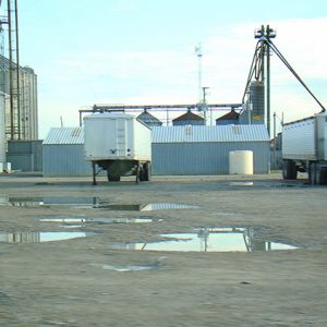 Semi-trucks and trailer parked outside large silos on parking lot with puddles