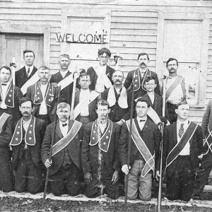 Group of white men in suits with different types of ribbons and sashes poses outside building