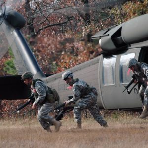 Three armed male soldiers hurriedly exiting powered helicopter in field near tree line