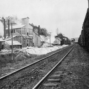 Railroad track running next to a two-story mining building with two smoke stacks and power lines