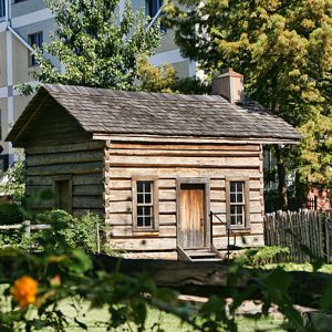 small log cabin in fenced-in plot with more modern city building, trees in background