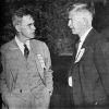 Two white man in suits lapel ribbons standing in casual conversation