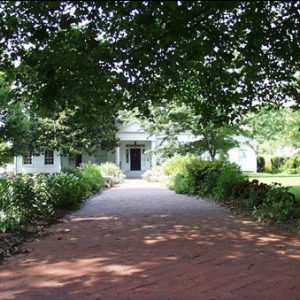 Wide brick sidewalk under tree-lined approach to white wood frame home