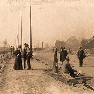 People standing in street and on sidewalks with rubble in vacant lots and tent next to destroyed building