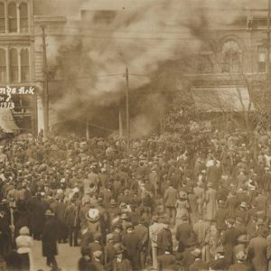 Large crowd of people outside watching city buildings burn