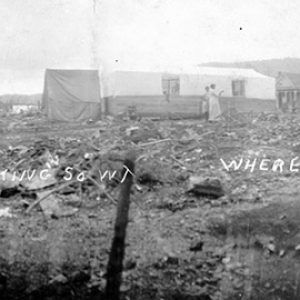 Debris covered lots with multistory house and men standing by tents after a fire